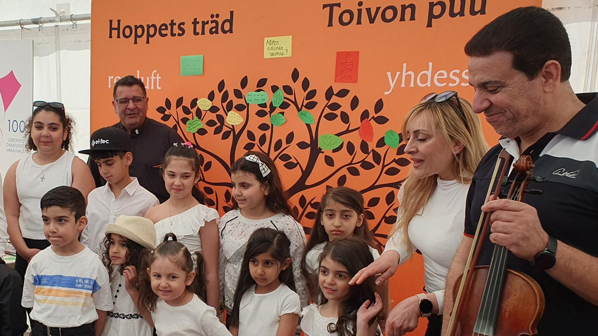 Lapsikuoro valkoisissa puseroissa kuoronjohtajan ja muusikoiden kanssa ryhmäkuvassa, taustalla piirretty toivon puu