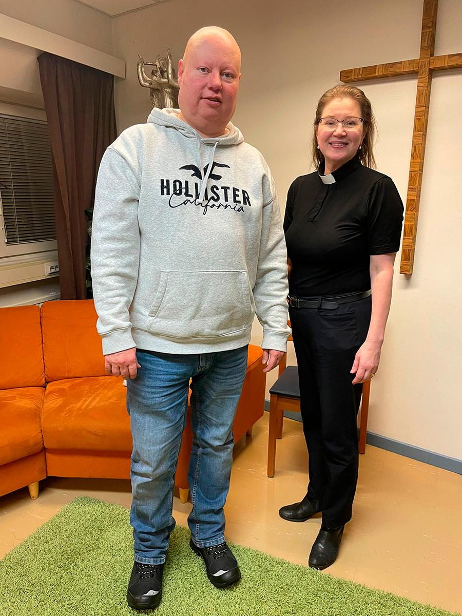 Petteri Auvinen ja Virpi Koskinen seisovat vierekkäin, taustalla oranssi sohva ja seinällä iso puinen risti.