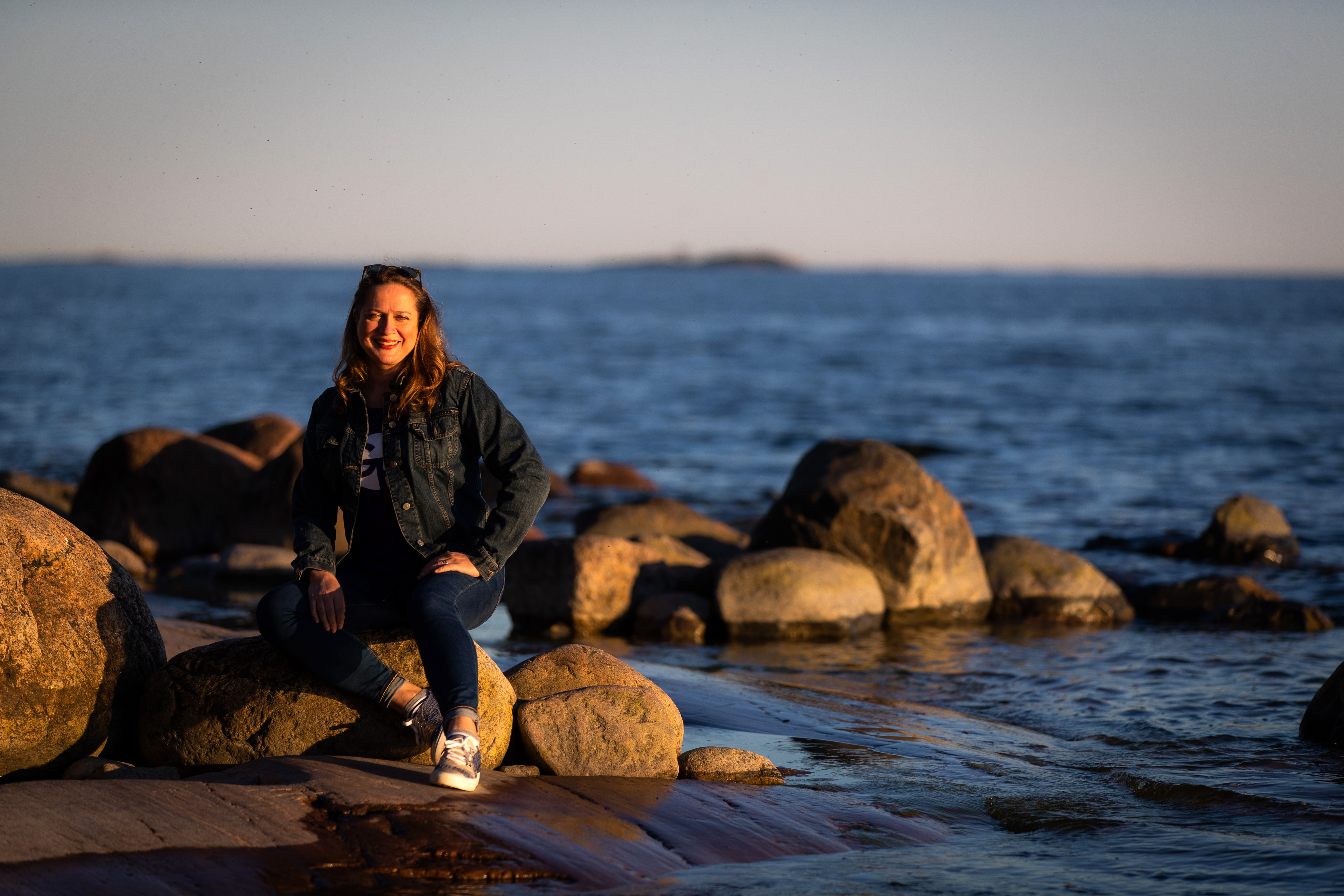 Hymyilevä pitkätukkainen nainen istumassa kiven päällä meren rannalla