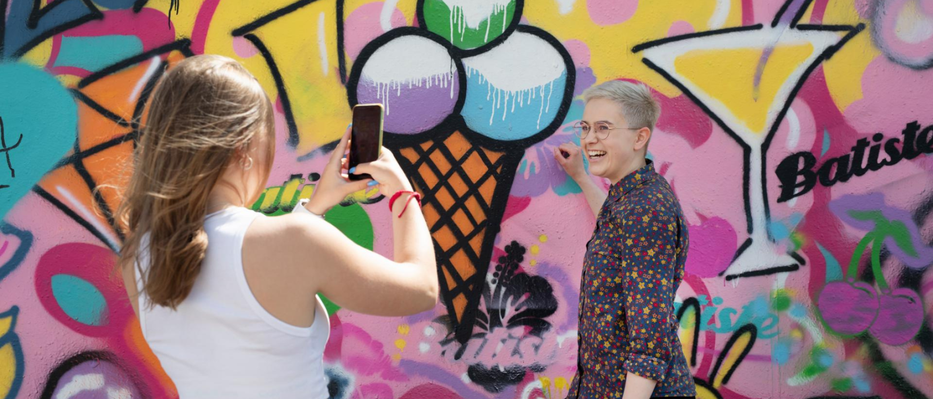 Nuori nainen ottaa valokuvaa toisesta naisesta, taustalla graffittiseinä
