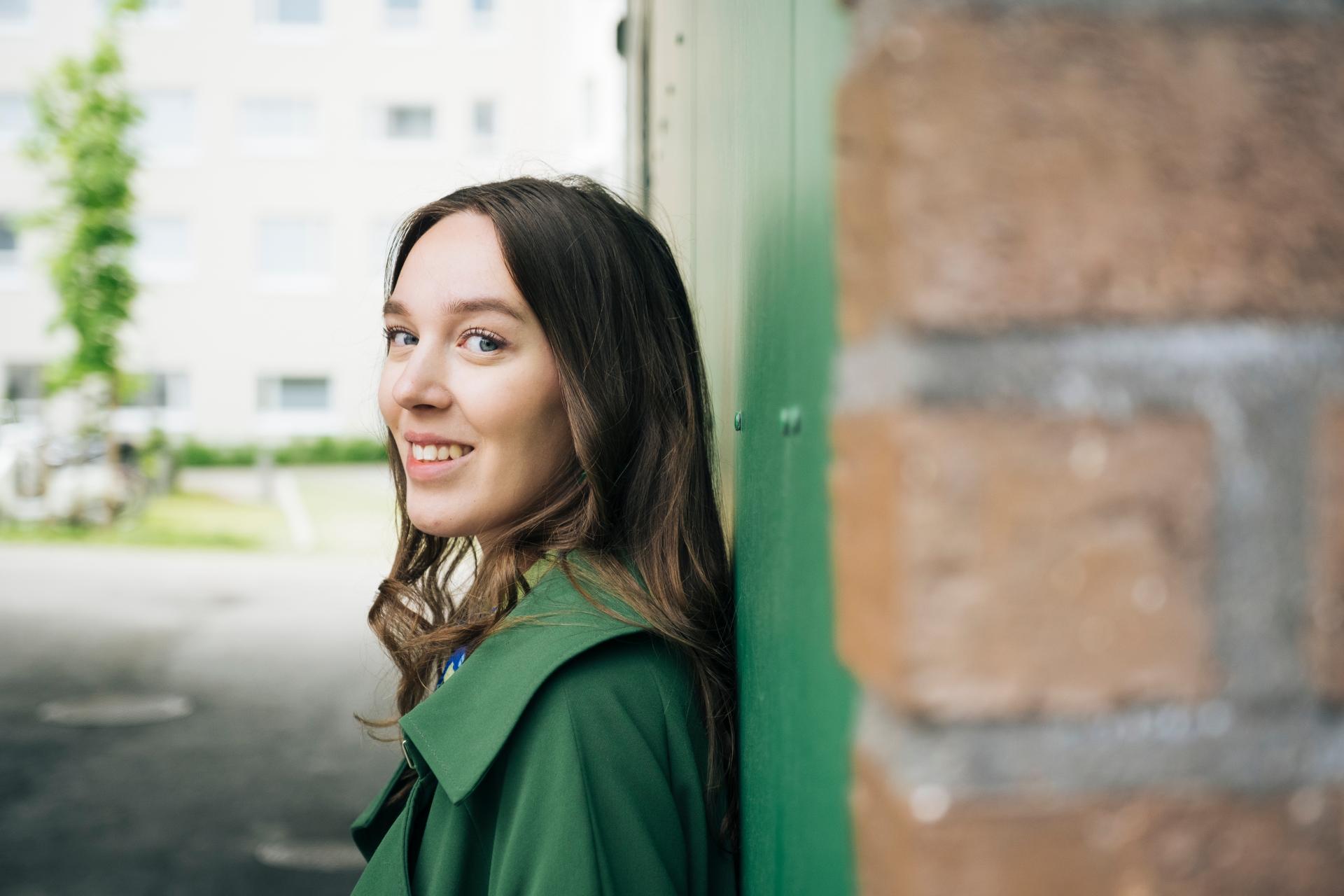 Nainen vihreässä takissa nojaa seinään ja katsoo kameraan.