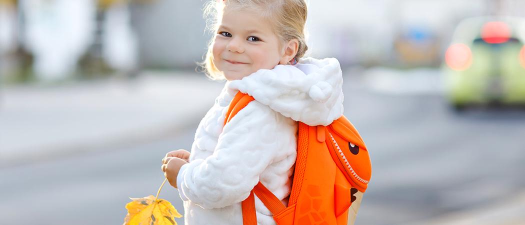 Pieni hymyilevä tyttö oranssi reppu selässään. Tytöllä kädessä syksyn keltaiseksi värittämä vaahteranlehti.