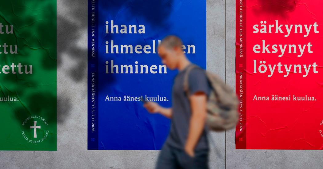 Nuori mies katsoo kännykkää samalla kun kävelee, taustalla seurakuntavaalien julisteita joissa lukee: ihana ihmeellinen ihminen