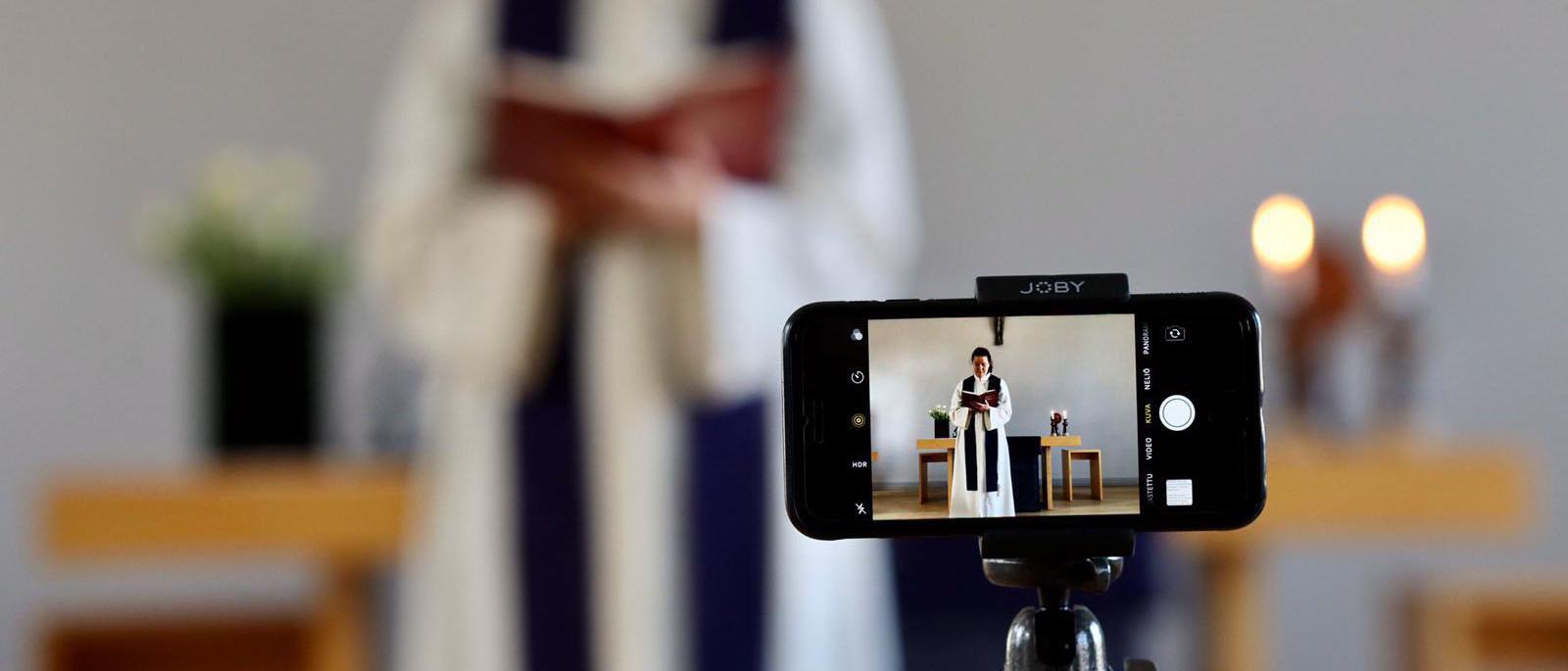 Kuvassa etualalla kännykkä alustalla, jolla kuvataan kirkossa pappia kasukassa.