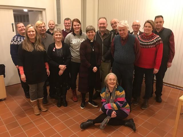 Pakilan seurakunnan seurakuntaneuvosto. 14 ihmistä seisoo rykelmässä, yksi nainen istuu heidän edessään lattialla.