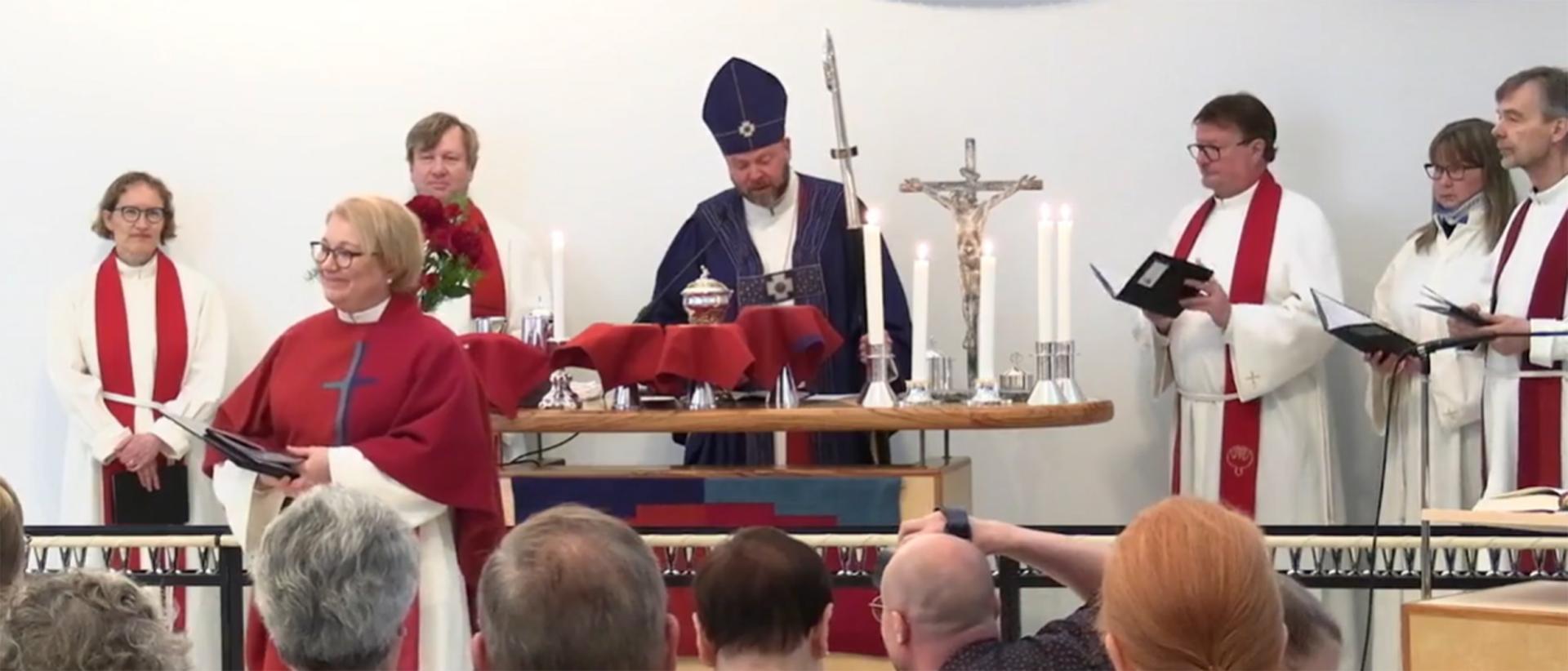 Kirkkoherra Minnamari Helasepän virkaansiunaaminen Myllypuron kirkossa 5.6.2022 Kuvassa piispa Teemu Laajasalo avustajineen ja  kirkkoherra polvistuneena selin. Kuvakaappaus videolta.