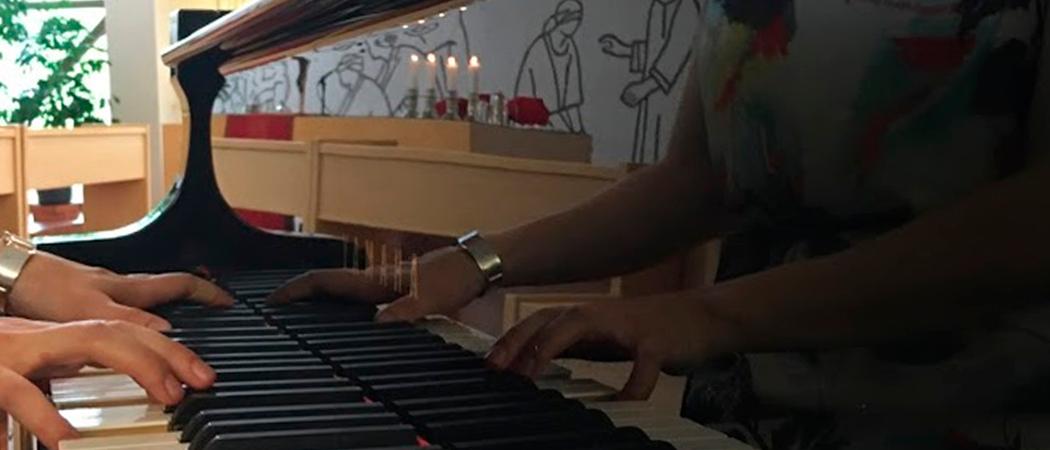 kuva Vuosaaren kirkon kirkkosalista, kanttori soittaa pianoa jonka pinnasta heijastuu kirkon alttaritaulun kuva ja alttarikynttilät