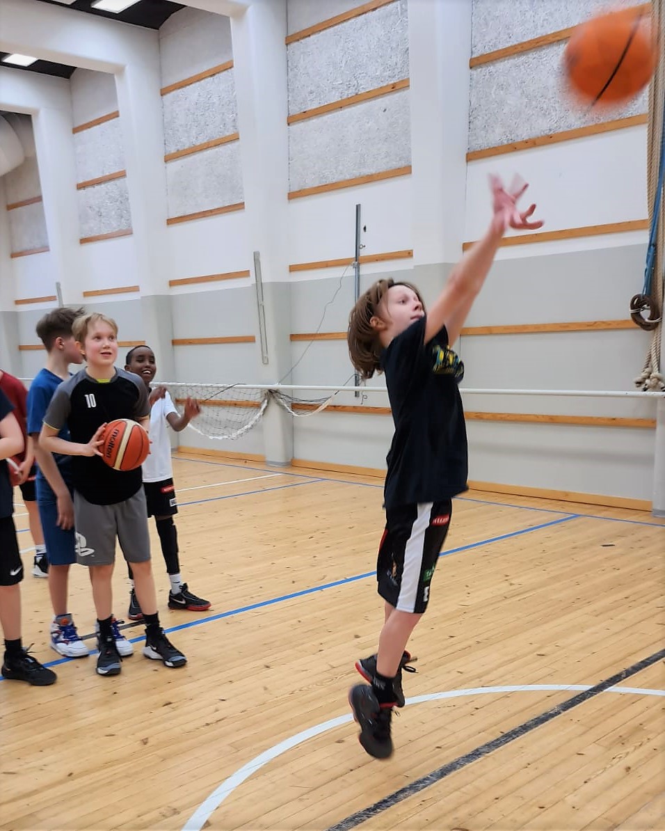 Alppila Basket koripallojoukkueen lapset harjoittelevat. Vasemmalla pojat odottavat vuoroaan, kun oikeanpuolimmainen poika heittää pallon koriin.
