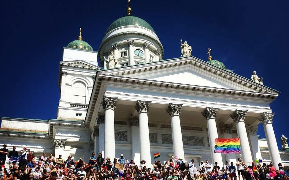 Tuomiokirkon portailla on paljon Helsinki Pride -kulkueeseen osallistuvia ihmisiä, sateenkaarilippuja, ilmapalloja.