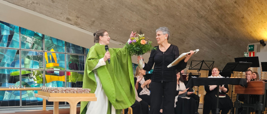 Kirkkoherra Virpi Koskinen ojentaa moniväristä kukkakimppua kanttori Anne Myllylälle Kannelmäen kirkossa. Taustalla Vokaaliyhtye CantoNovon laulajia.