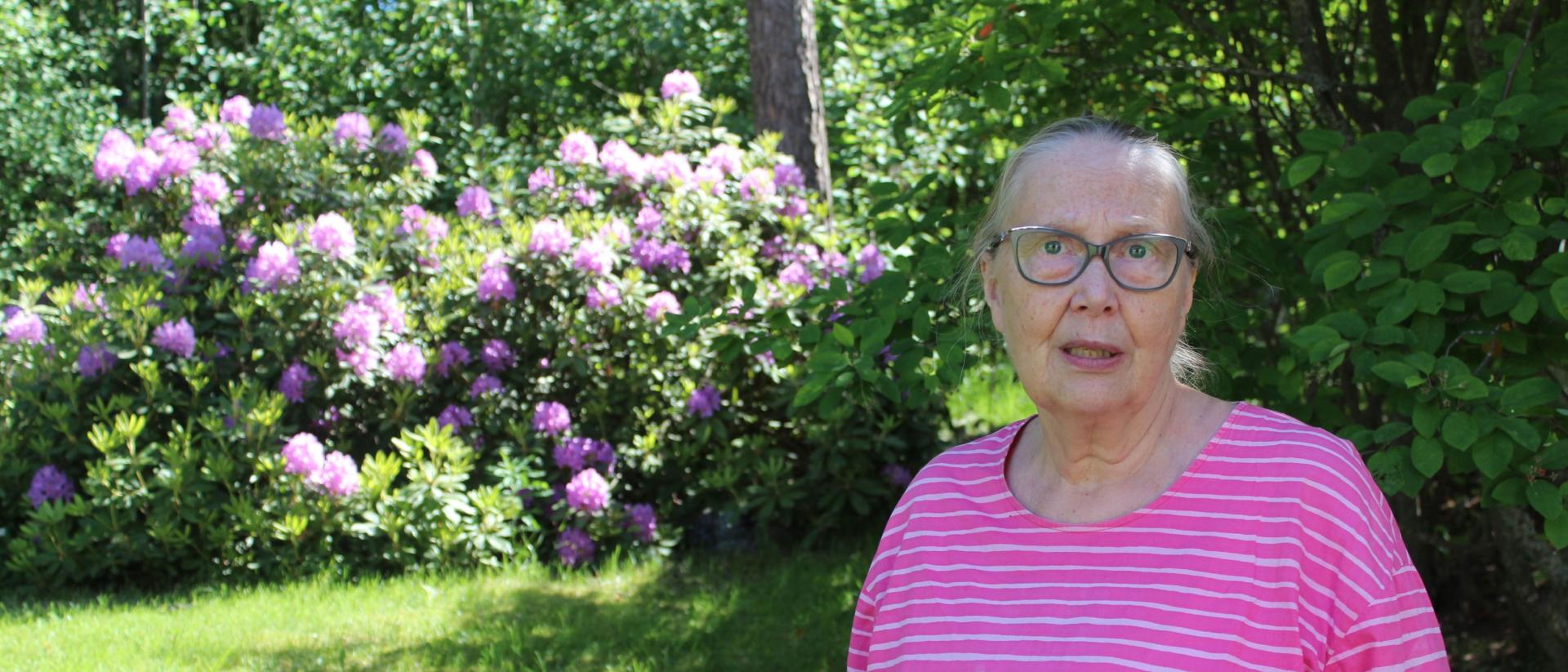 Mikaelin seurakunnan pastori Marja Lehtinen kuvattu kirkon takapihalla, taustalla violetti alppiruusu.