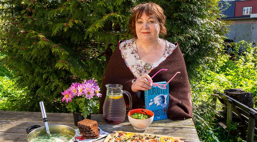 Kirjailija Imbi Paju kuvattuna kesäisessä puutarhassa ruokapöydän ääressä kirja kädessä- Kuva: Sanna Larmola