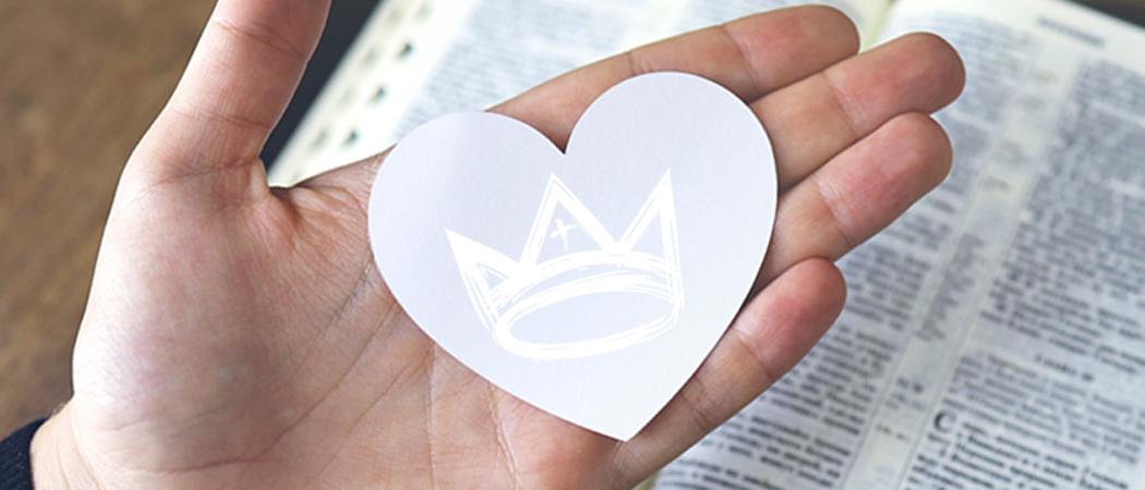 Avoinaisella kämmenellä paperisydän, jossa Kuninkaan iltojen tunnus, kruunu. Taustalla avoinainen Raamattu.