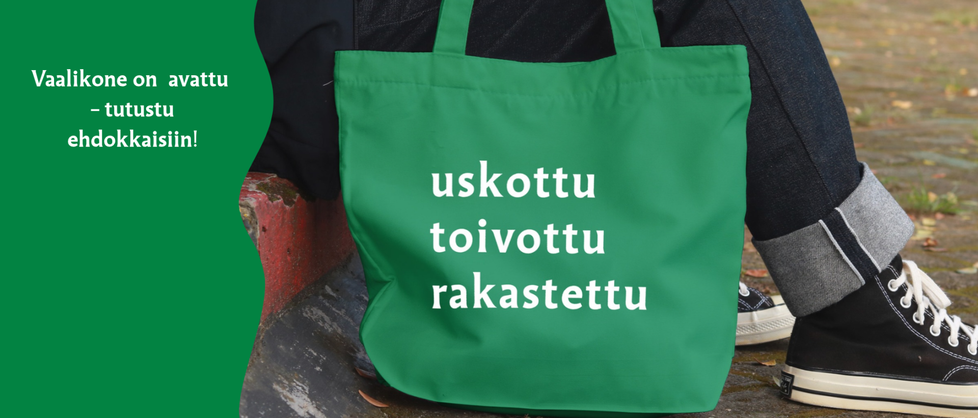 Maassa vihre&auml; kassi, jossa teksti uskottu toivottu rakastettu. Kassia pitelee henkil&ouml;, jolla tennarit.