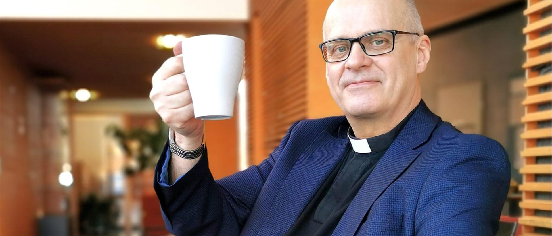 Roihuvuoren srk pappi Kari Laaksonen kohottaa kahvikuppia Laajasalon kirkon aulassa tumma puku yllään. Kuva: Marko Anttila