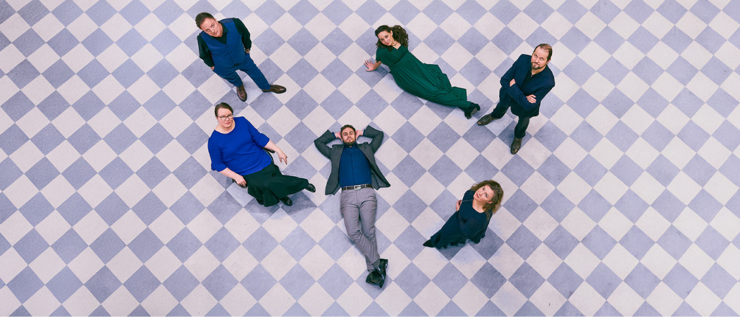 The Vicentino Singers -lauluryhm&auml;n kuusi j&auml;sent&auml; violettien v&auml;risell&auml; laattalattialla katsoo yl&ouml;s kameraan.