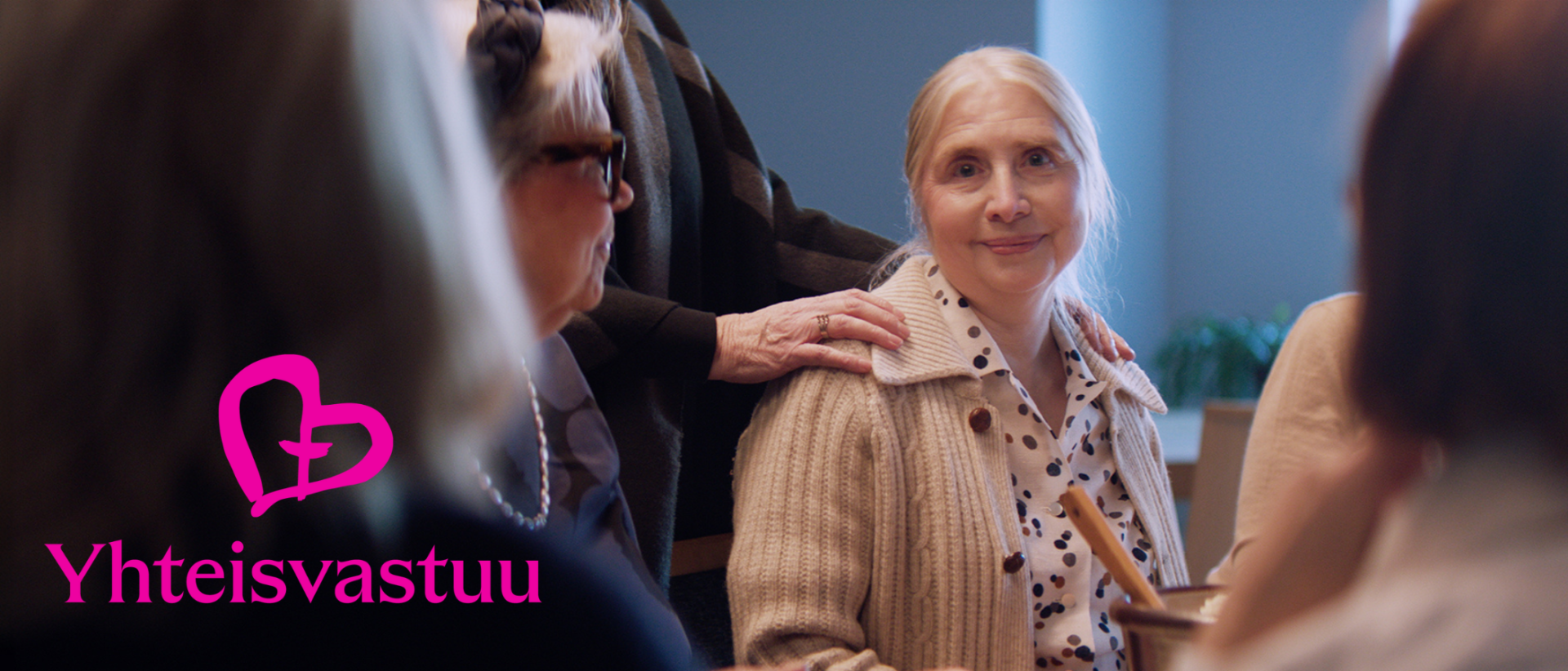 Nainen, jolla beige villatakki, istuu ja katsoo kameraan, toisen naisen k&auml;si olkap&auml;&auml;ll&auml;&auml;n.