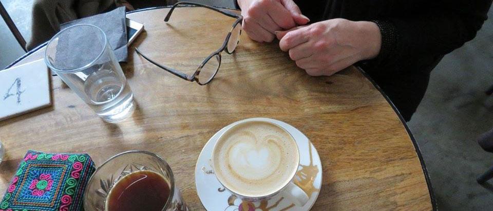 Kuvassa pöydän ääressä istuu ihminen ja pöydällä kahvikuppi ja silmälasit