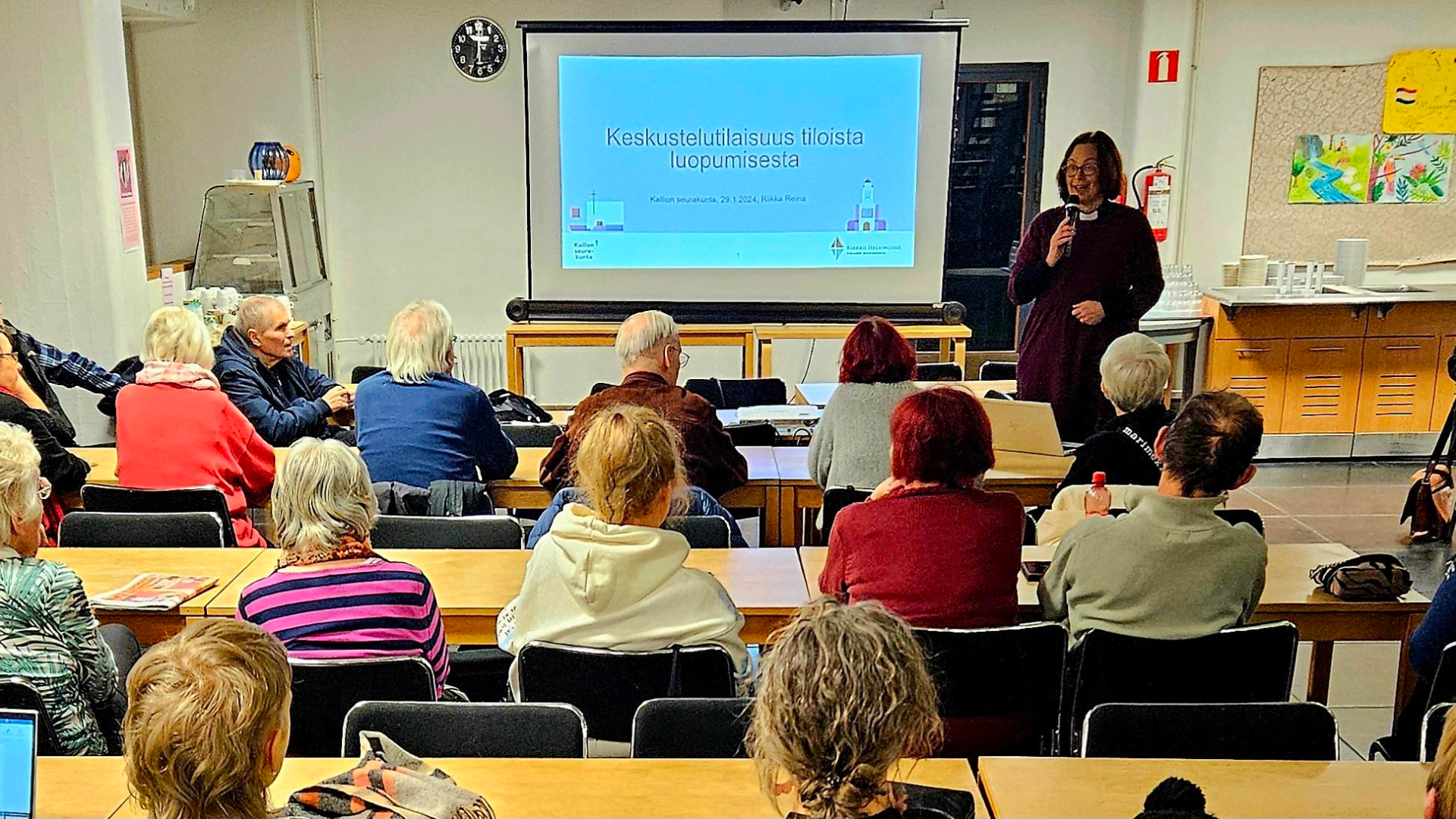 Edessä kalvo, oikealla Riikka Reina, yleisöä istumassa selin