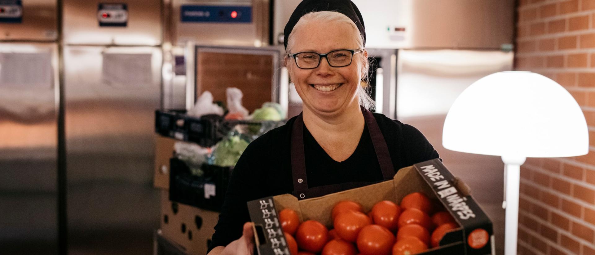 Hymyilevä nainen kokin asussa katsoo kameraan tomaattilaatikko edessään