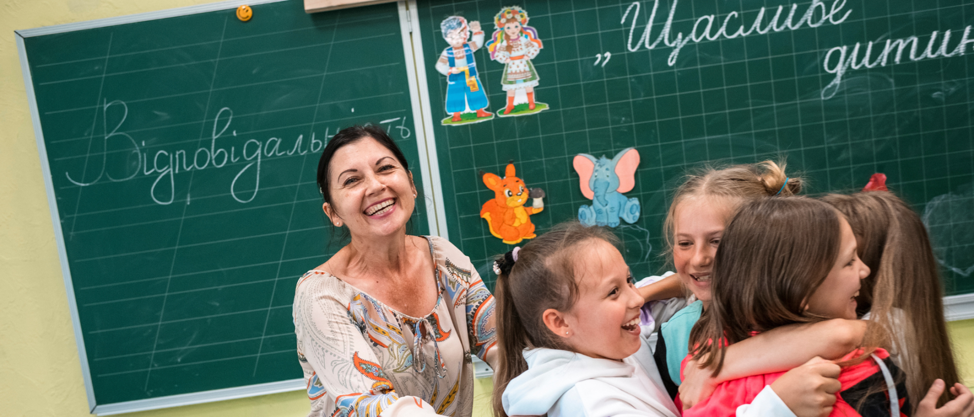 Opettaja ja joukko lapsia koululuokassa, taustalla liitutaululla kirjoitusta ukrainan kielellä