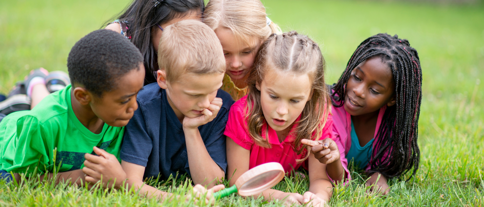 Lapsia vatsallaan nurmikolla katsomassa suurennuslasilla jotain