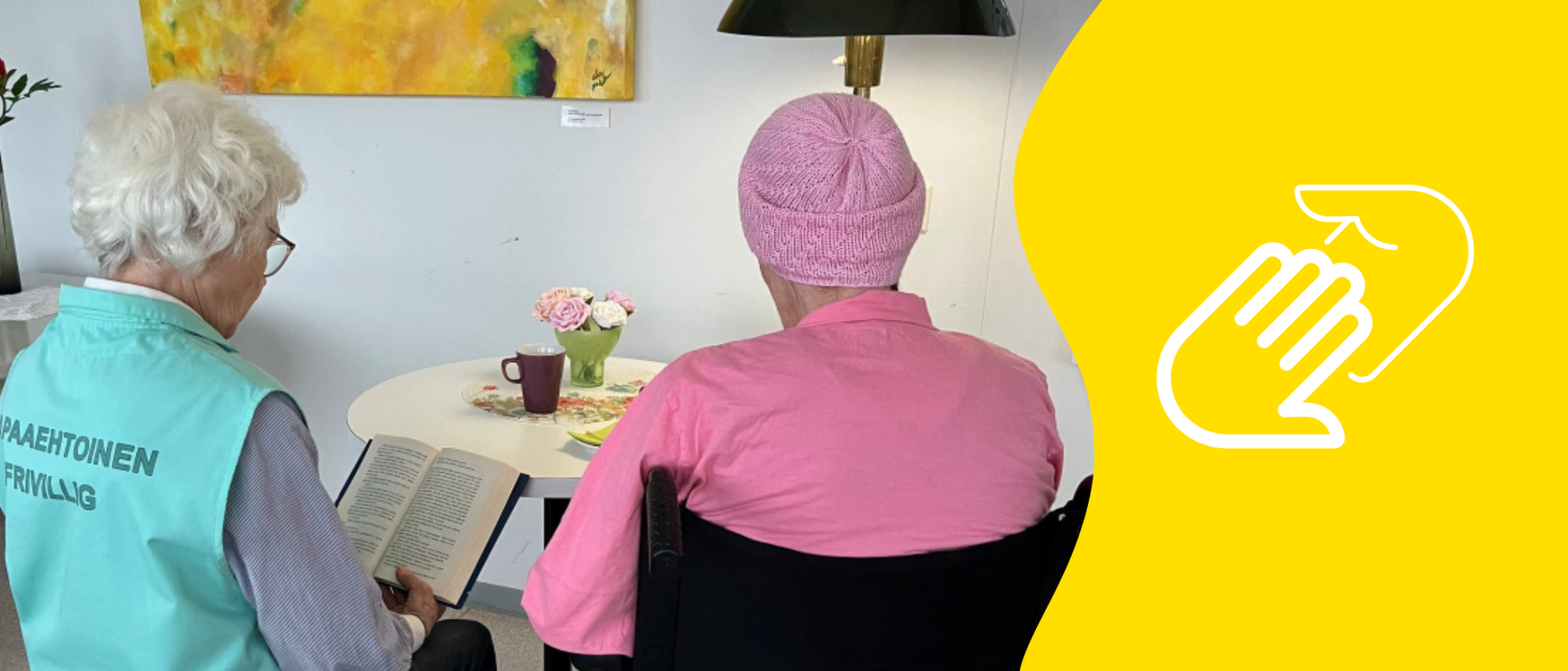 Kaksi naista istuu selin kameraan, vasemmanpuoleisen selässä lukee vapaaehtoinen ja oikeanpuoleinen istuu pyörätuolissa.