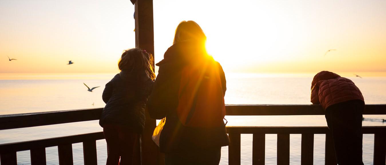Aikuinen ja kaksi lasta katselevat merelle auringonlaskua.
