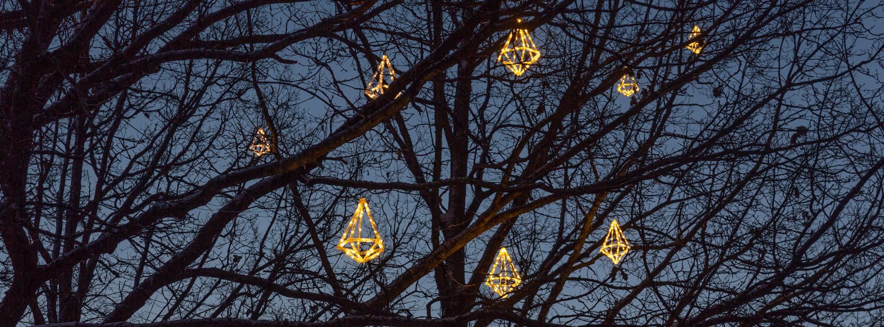 Hämärässä loistavia, puun oksistoon ripustettuja timantin muotoisia jouluvaloja.