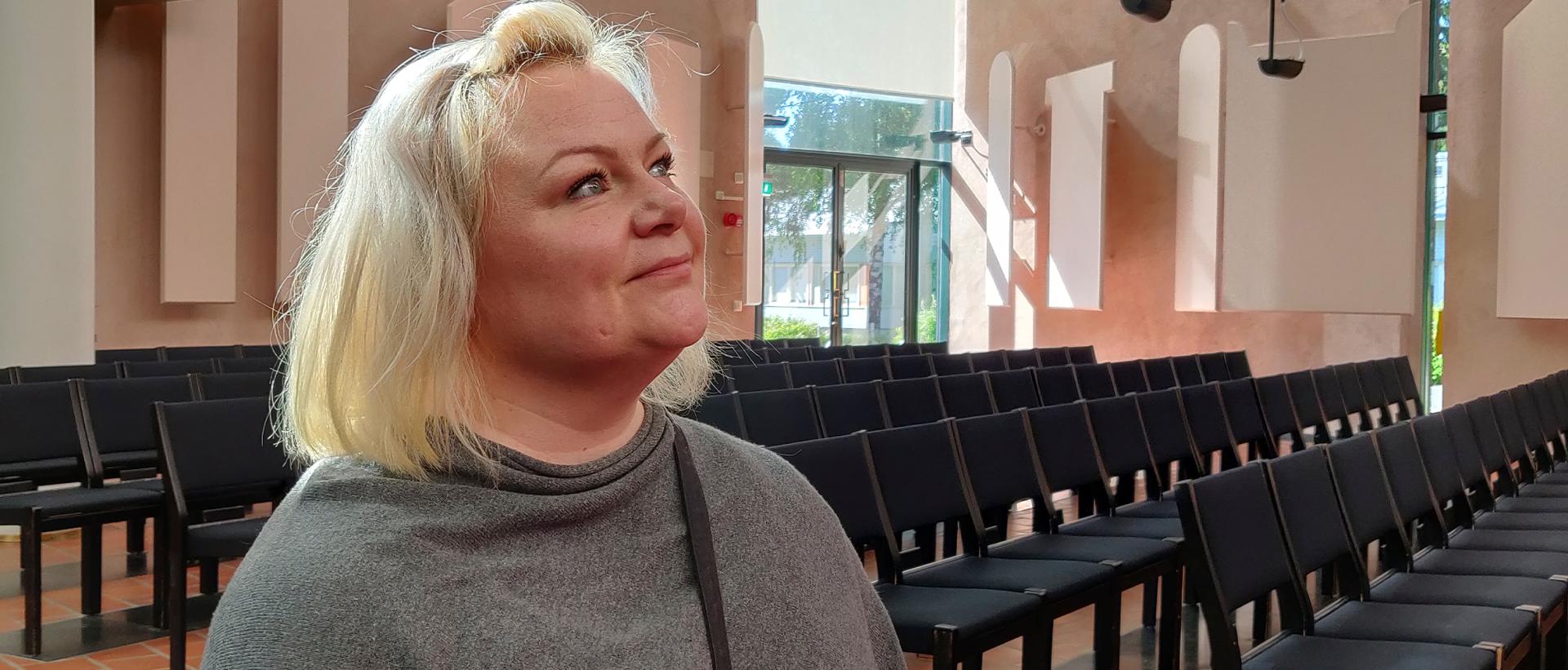 Sini Ikävalko istuu Mikaelinkirkon penkissä katse yläviistoon, taustalla lasiovi luontoon