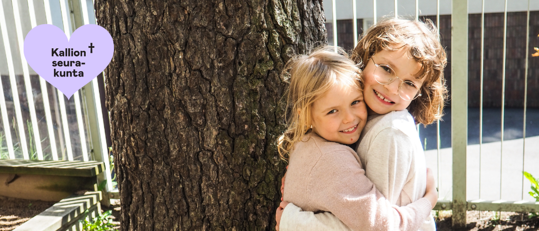 Kaksi lasta halaa toisiaan puun luona hymyillen