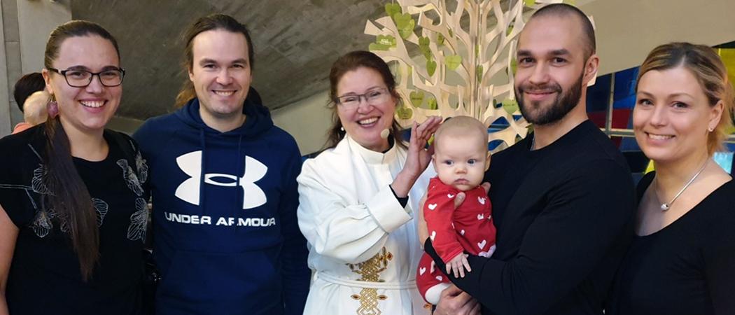 Kannelmäen kirkossa kastepuun edessä kaksi pariskuntaa vauvakirkkoon osallistuneita vanhempia ja kummeja papin vieressä. Keskellä vauva punaisessa vaatteessa.