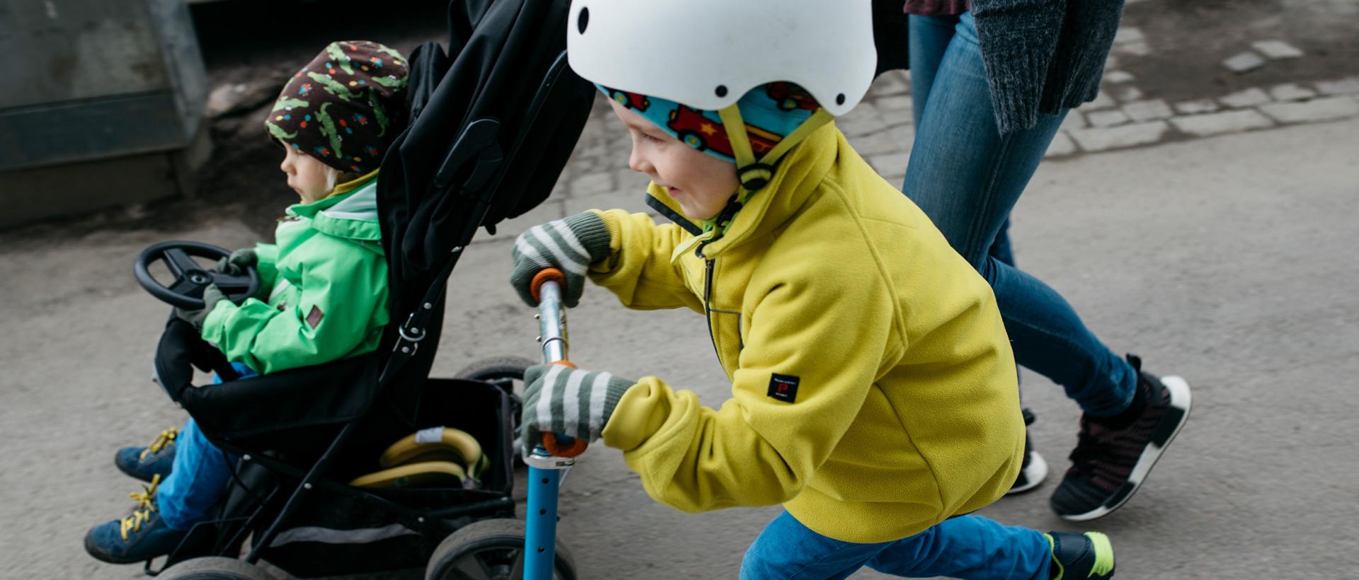 Två barn, en i vagn och en på sparkcykel. En förälder skuffar vagnen.