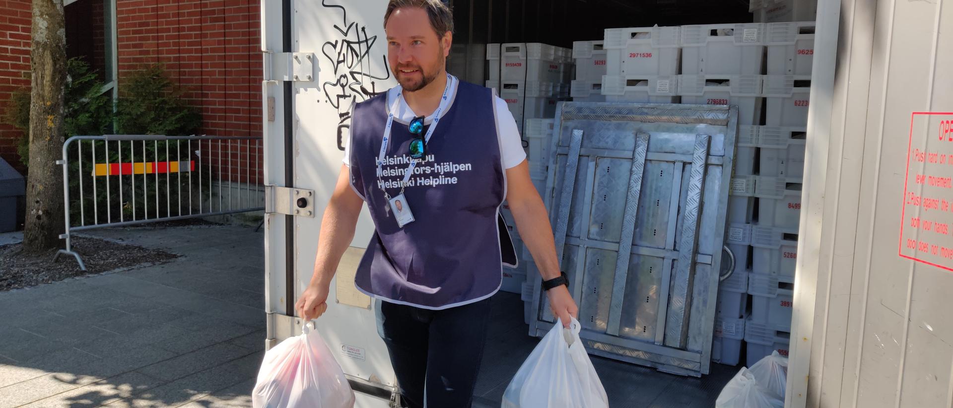 Mies, jolla on yllään Helsinki-avun liivi, kantaa kahta valkoista muovikassia täynnä ruokaa.