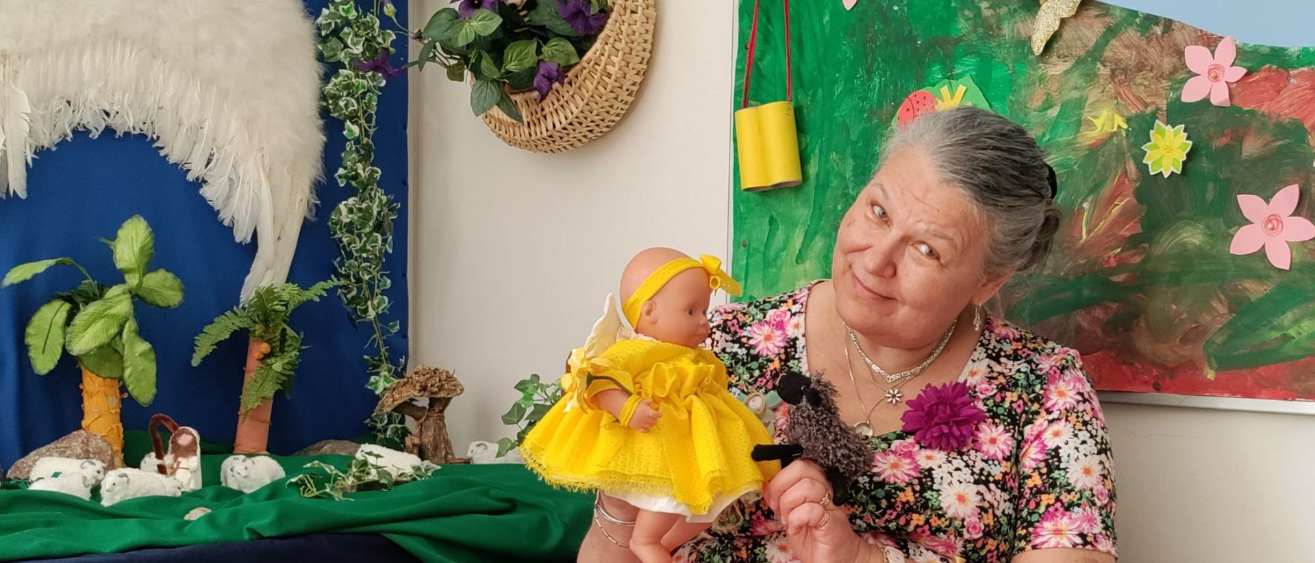 Mikaelin seurakunnan lastenohjaaja Auli Karhu päiväkerhotilassa kädessään keltaisessa mekossa oleva nukke ja lammas
