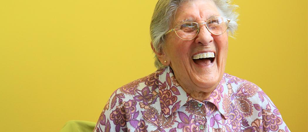 Iloisesti naurava iäkäs nainen keltaista taustaa vasten.