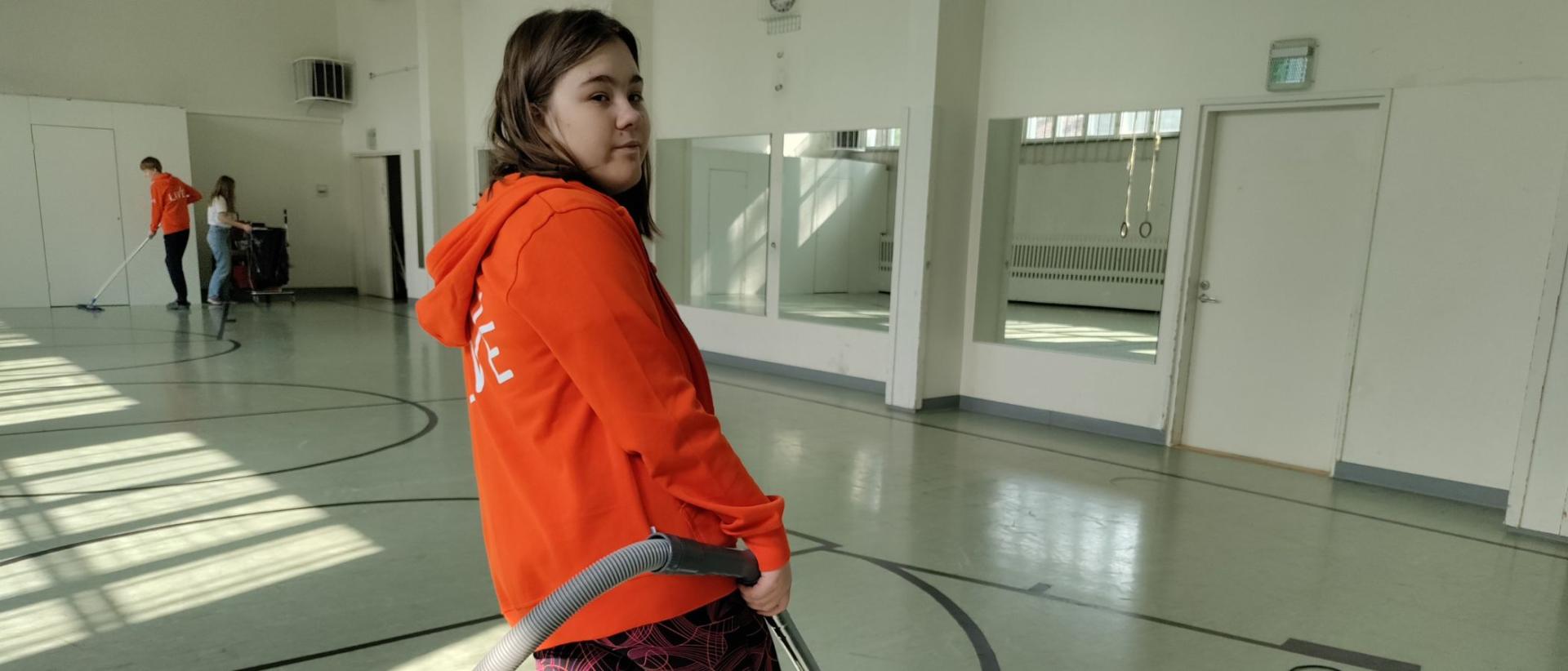 TELMA-opiskelija Hertta siivoamassa Vartuiokylän kirkon jumppasalia imuri kädessä oranssissa hupparissa