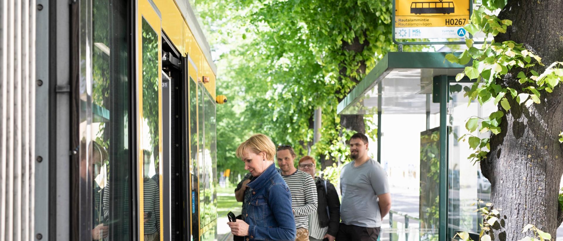 Ihmisiä ratikkapysäkilläkesäaikaan, nainen nousee juuri ratikkaan