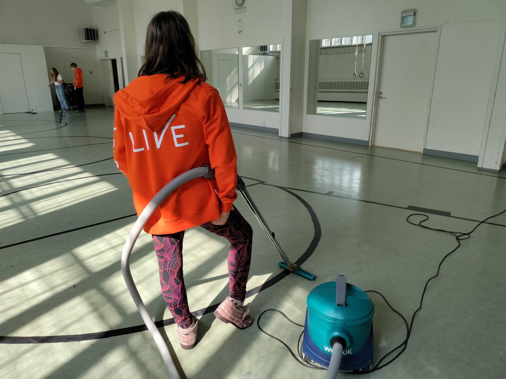 TELMA-opiskelija Hertta siivoamassa Vartiokylän kirkon jumppasalia selin, oranssissa hupparissa luke valkoisella LIVE