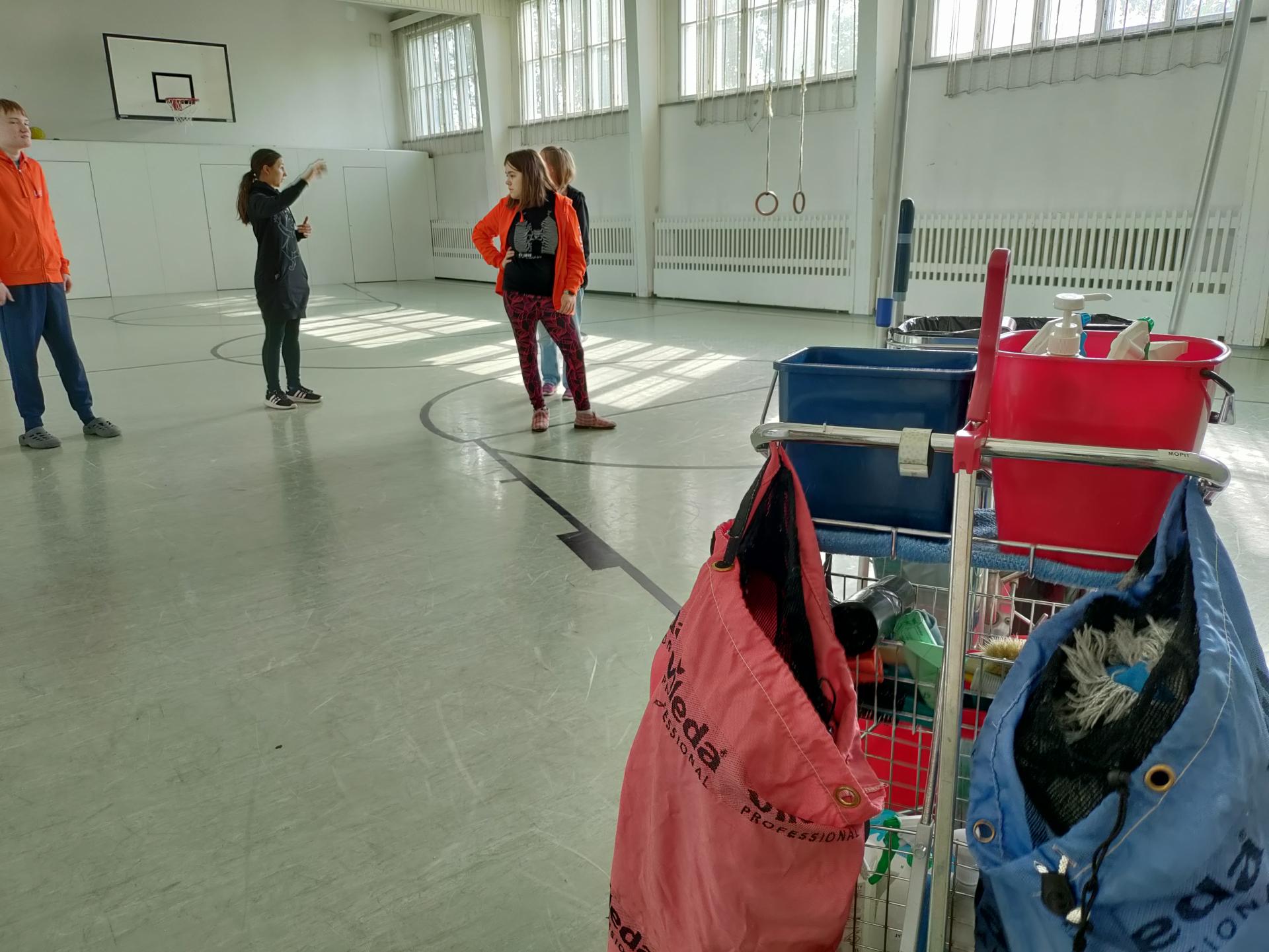 Siivouskärry Vartiokylän kirkon jumppasalissa. Taustalla TELMA-opiskelijoita ja ohjaaja