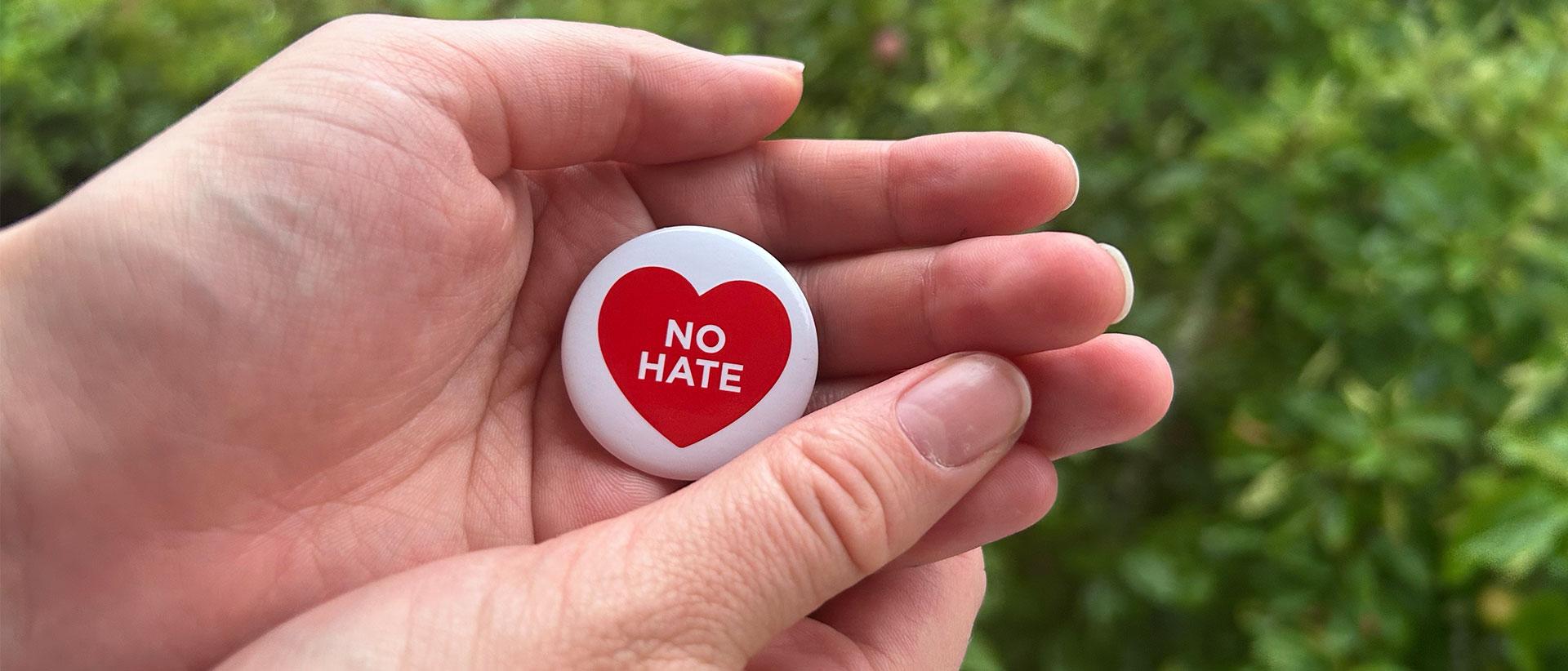 Kädessä rintanappi jossa valkoisessa pohjalla punainen sydän tekstillä no hate