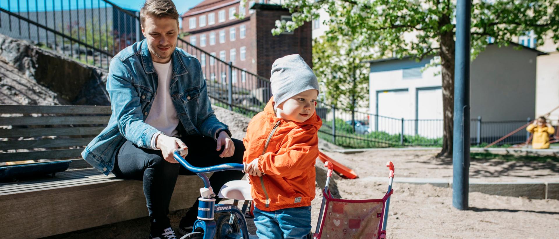Lapsella oranssi huppari ja sininen polkupyörä, taustalla isä pitää kiinni pyörästä