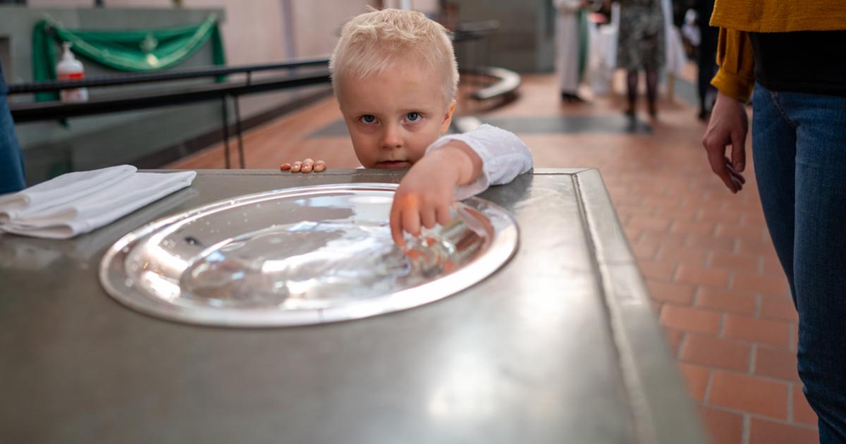 Pieni vaaleatukkainen poika kokeilee kastemaljan vettä sormillaan MIkaelinkirkon kastepäivässä. Kuva: Heikki Heikurinen