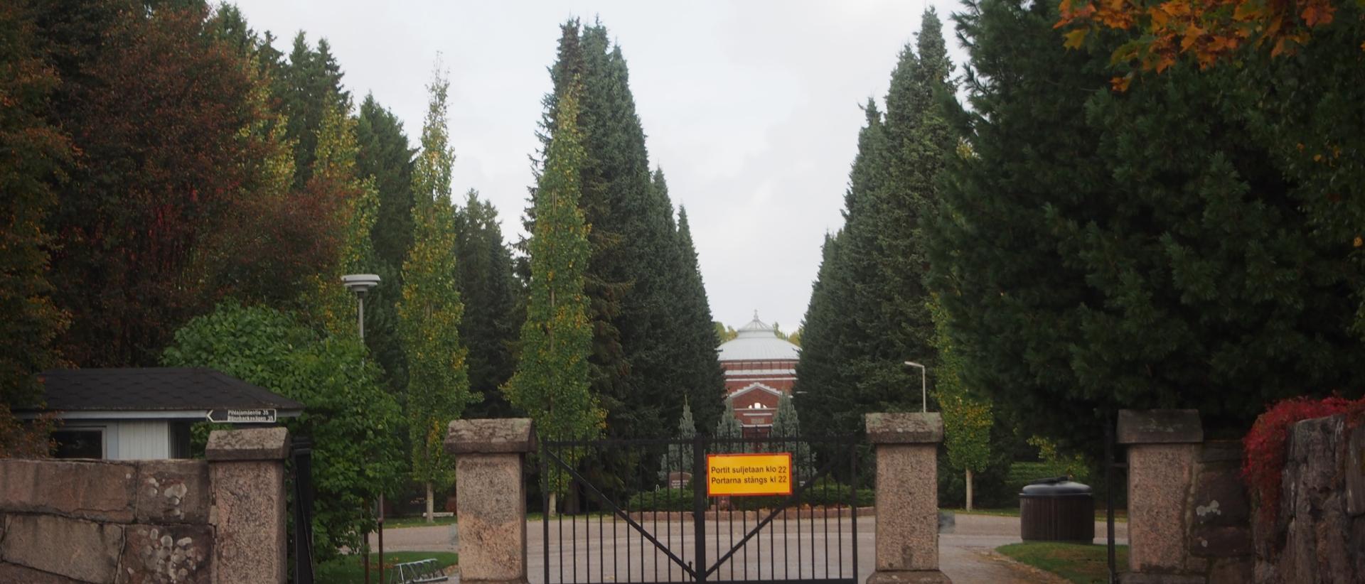Etualalla rauta-aita ja suljettu portti, taaempana puita ja pensaita sek&auml; n&auml;kym&auml; Malmin hautausmaan kappelille