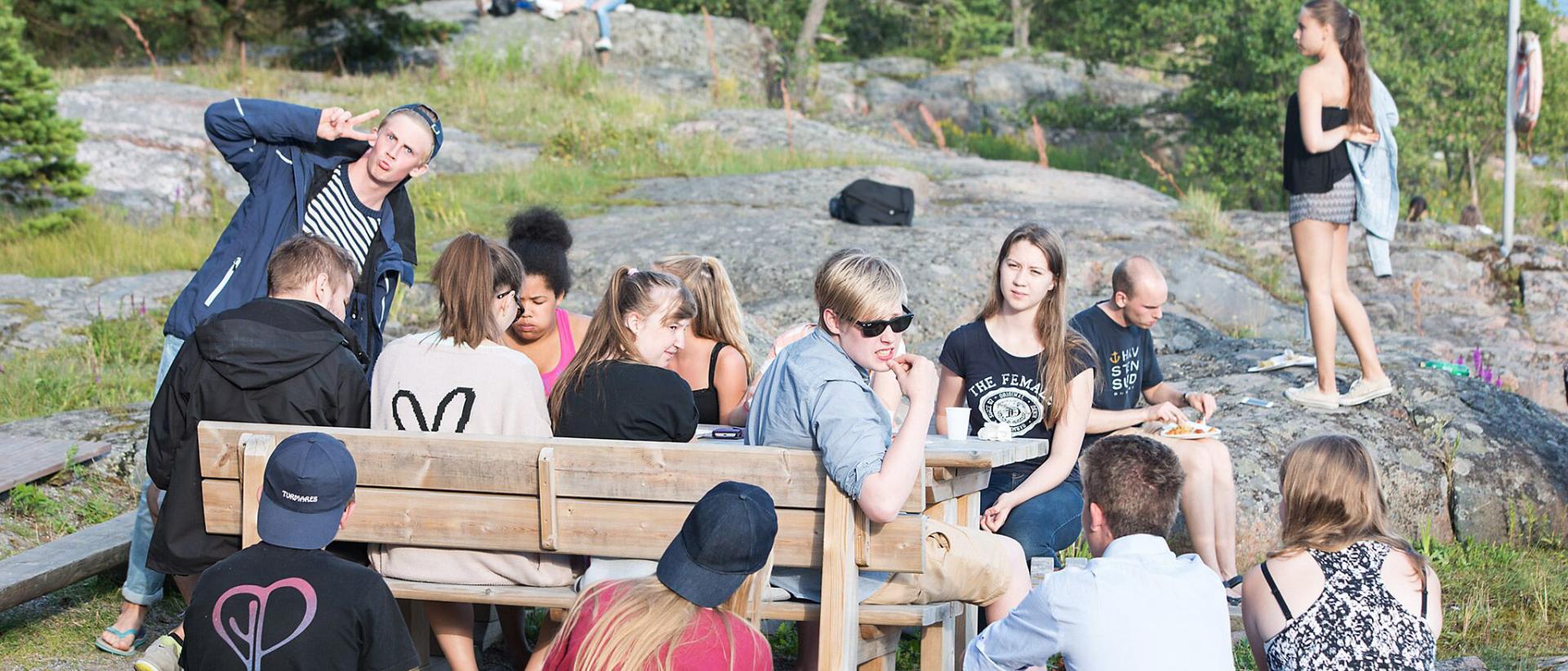 Ungdomar sitter på bänkar vid klipporna ute på Lekholmen. De äter lunch.